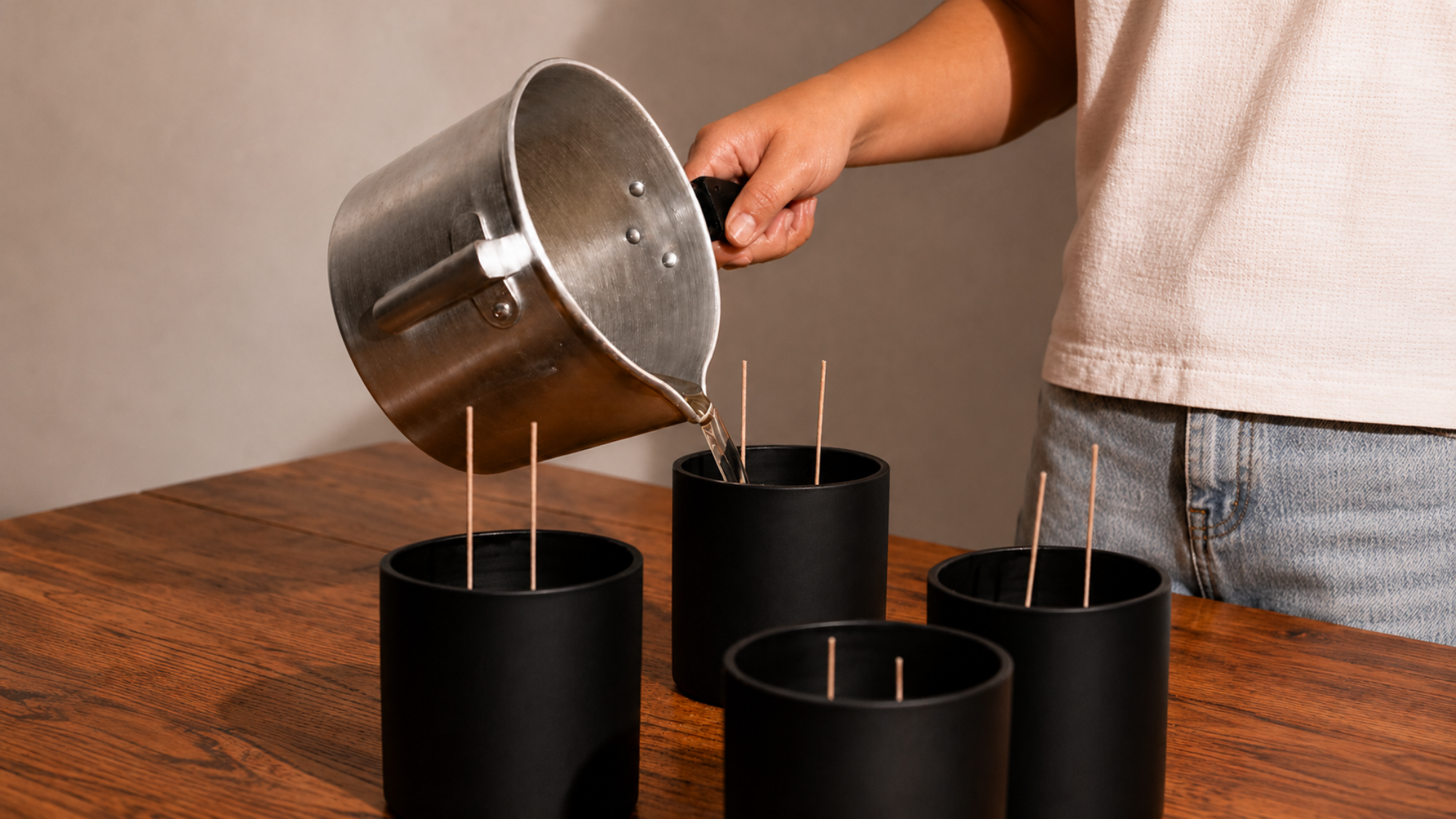 Person pouring liquid from a pot into black containers with sticks on a wooden table.