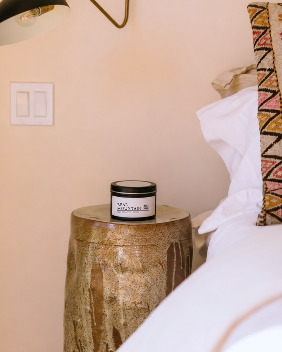 A Bear Mountain candle in a black tin with a white label sits on a rustic stone side table next to a bed with white linens and a patterned pillow in a warm beige bedroom setting.
