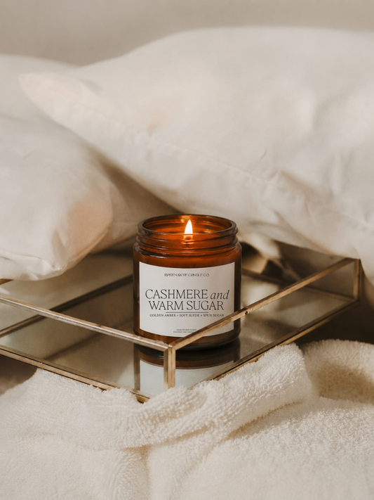 Candle labeled 'Cashmere and Warm Sugar' on a metallic tray with pillows and blanket in the background