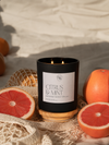 A black candle jar with a white label reading 'Citrus & Mint' sits on a wooden coaster, surrounded by citrus fruits and cream-colored fabric in a styled setting.