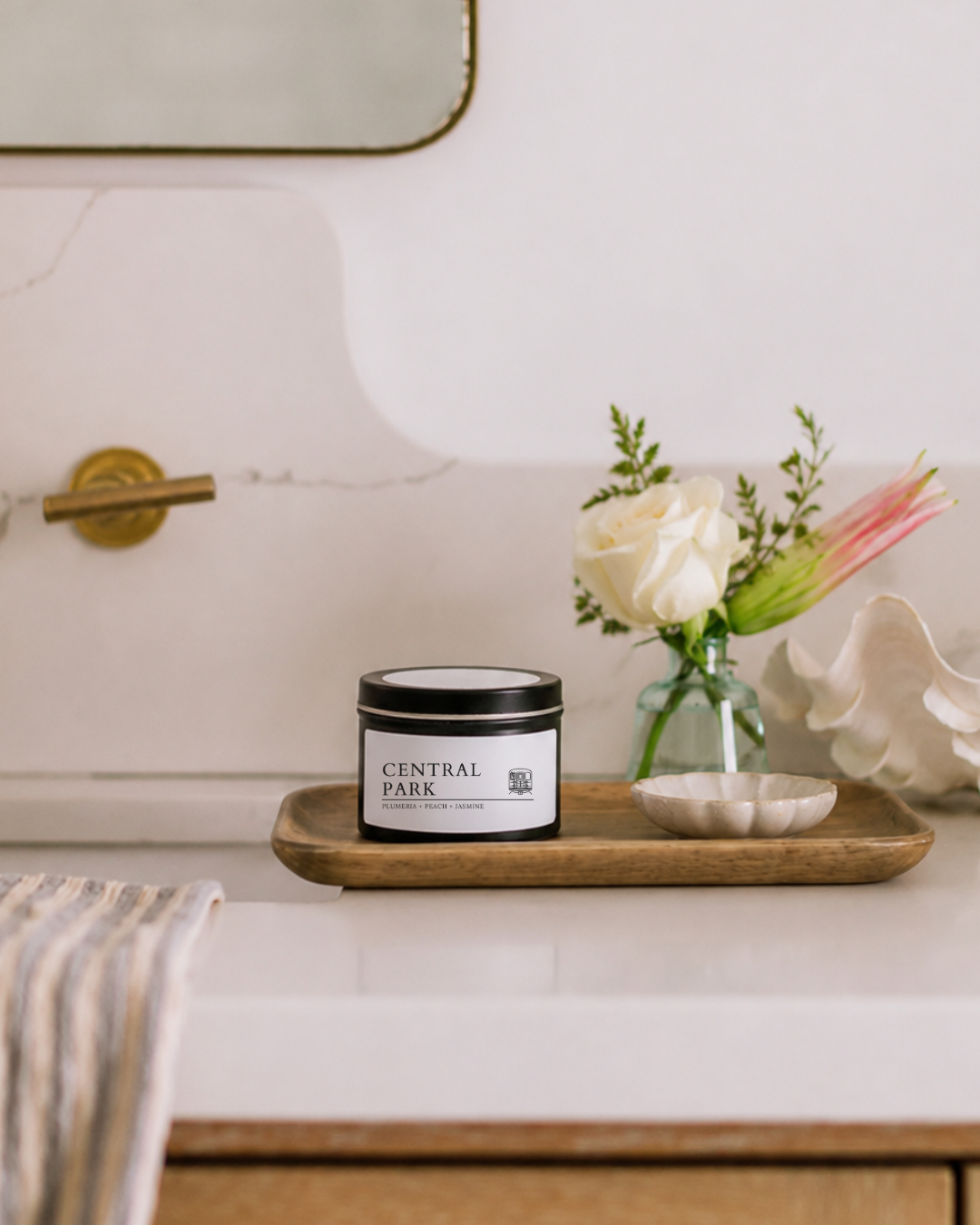 A candle tin labeled CENTRAL PARK sits on a wooden tray on a bathroom counter, styled with a small vase of flowers and a seashell.