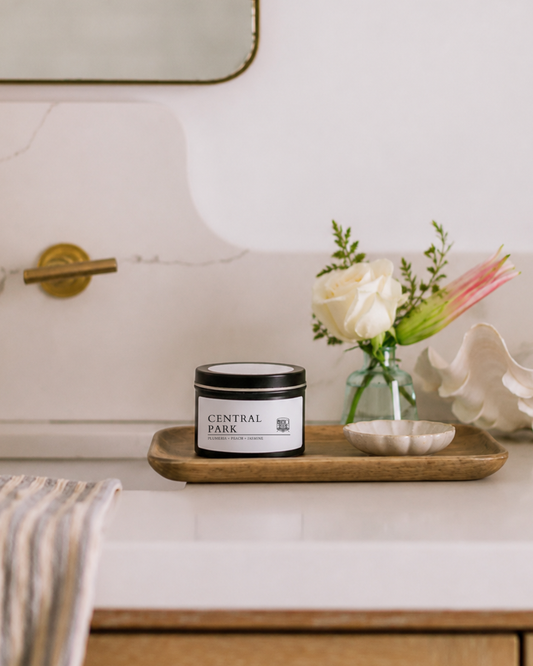 A candle tin labeled CENTRAL PARK sits on a wooden tray on a bathroom counter, styled with a small vase of flowers and a seashell.