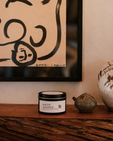 A black metal candle tin labeled 'FIFTH AVENUE' sits on a rustic wooden shelf beside a small ceramic object, with a framed abstract print on the wall behind it.