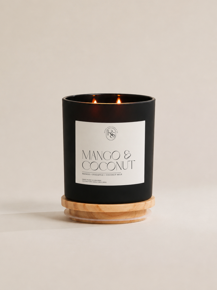 A black double-wick candle in a matte black glass jar with a white label reading 'MANGO & COCONUT' and a light natural wood base, photographed on a warm off-white background.