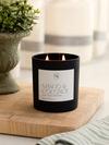 A black double-wick candle jar labeled 'MANGO & COCONUT' sits on a wooden board, with a blurred potted plant and striped cloth in the background.