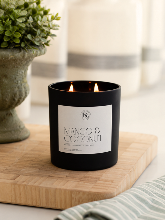A black double-wick candle jar labeled 'MANGO & COCONUT' sits on a wooden board, with a blurred potted plant and striped cloth in the background.