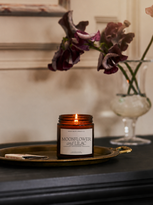 Candle labeled 'Moonflower and Lilac' on a gold tray with flowers in the background
