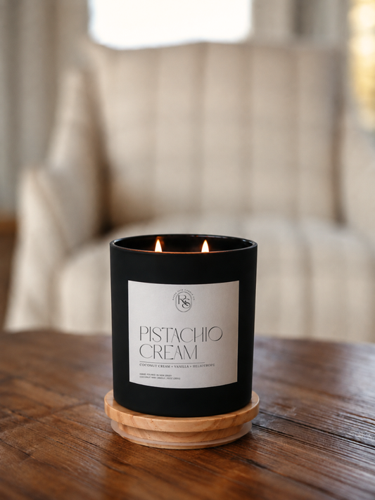 A black candle jar with a white label reading 'PISTACHIO CREAM' sits on a wooden coaster on a wooden table, with a softly blurred armchair in the background.