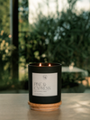 A black candle jar with two lit flames sits on a wooden coaster on a tabletop, with a white label reading 'PINE & CYPRESS' and a blurred outdoor garden background.