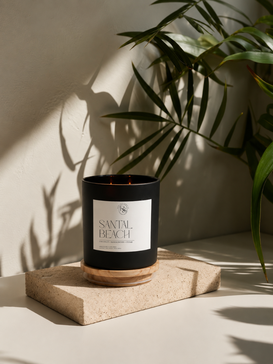 A black candle jar with a white label reading 'SANTAL BEACH' sits on a textured stone block, with warm sunlight casting leaf shadows on a beige wall and plant leaves in the background.