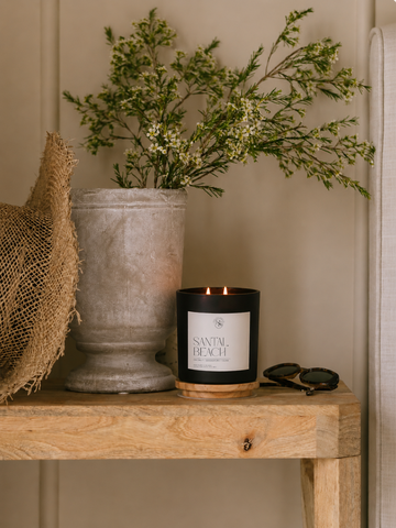 Candle with a label 'SANTAL BEACH' on a wooden surface next to a vase with greenery