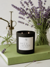A black candle jar labeled 'SAGE & LAVENDER' sits on a light green book, with lavender stems and a vintage-style brass pump bottle in the softly blurred background.