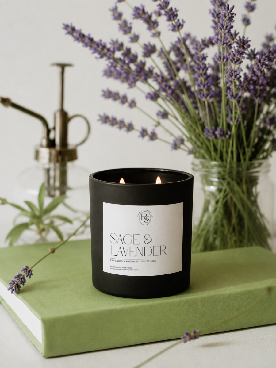 A black candle jar labeled 'SAGE & LAVENDER' sits on a light green book, with lavender stems and a vintage-style brass pump bottle in the softly blurred background.