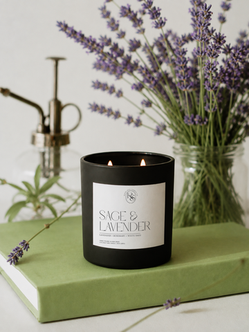 A black candle jar labeled 'SAGE & LAVENDER' sits on a light green book, with lavender stems and a vintage-style brass pump bottle in the softly blurred background.