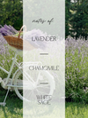 A vertical graphic with a faded lavender field and a white bicycle carrying a basket of purple lavender. Text in the center reads 'notes of: LAVENDER — CHAMOMILE — WHITE SAGE'.