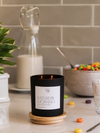 A matte black candle jar with a white label reading 'SATURDAY MORNING' sits on a wooden coaster on a kitchen counter, with a glass milk bottle, bowl of cereal, and tiled backsplash in the background.