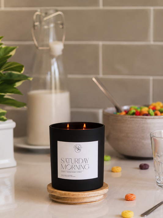 A matte black candle jar with a white label reading 'SATURDAY MORNING' sits on a wooden coaster on a kitchen counter, with a glass milk bottle, bowl of cereal, and tiled backsplash in the background.