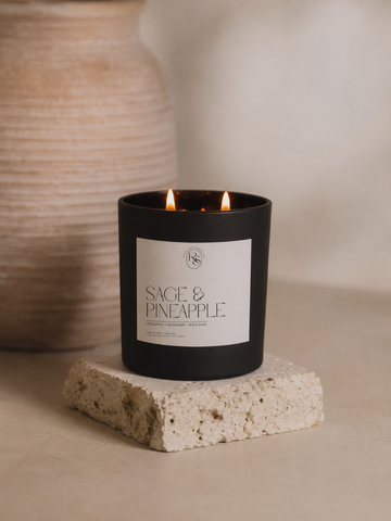 A black double-wick candle labeled 'SAGE & PINEAPPLE' sits lit on a textured stone block, with a warm beige ceramic vase blurred in the background.