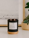 A black candle jar with a white label reading 'SEA SALT & CEDAR' sits on a round wooden coaster on a tabletop. The candle is lit, with two flames visible, and is styled in a home setting next to a potted succulent and a framed black-and-white ocean print.
