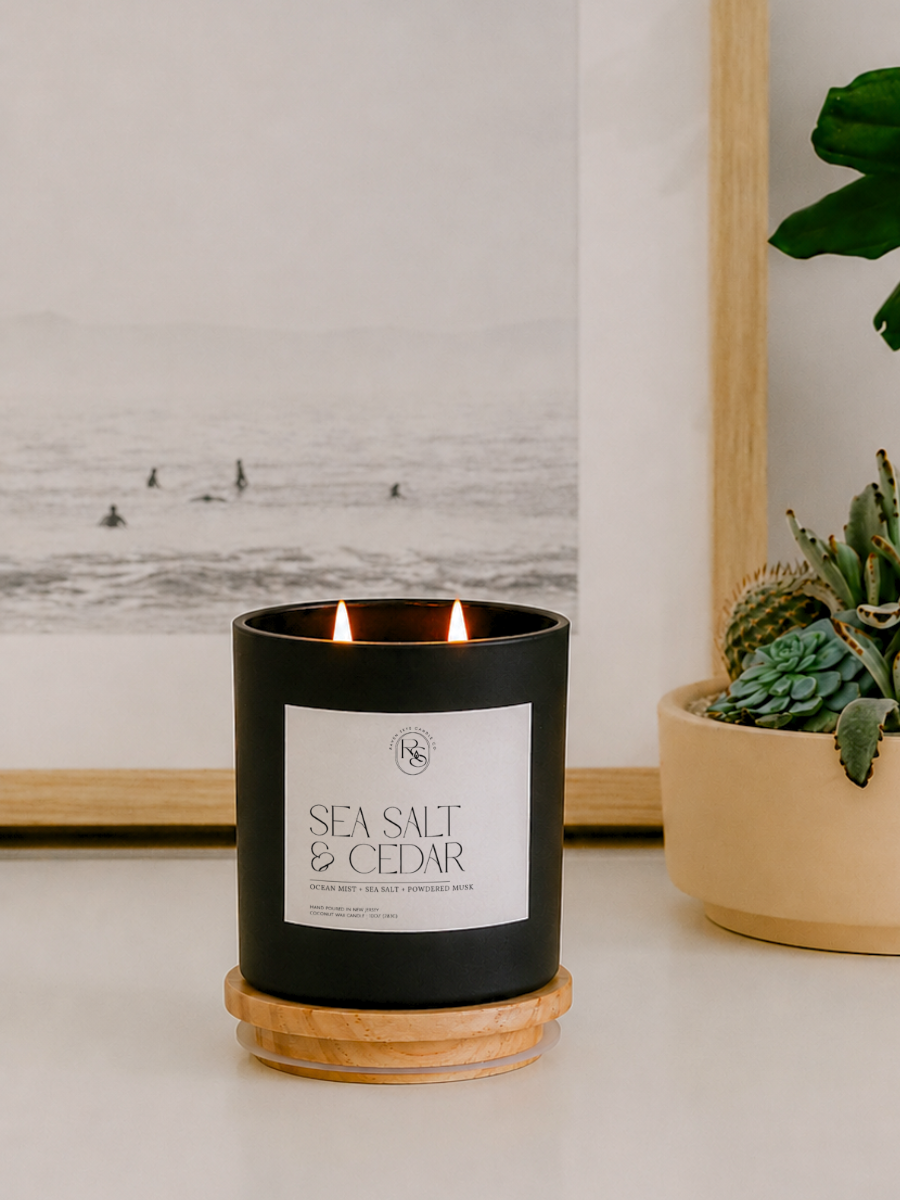 A black candle jar with a white label reading 'SEA SALT & CEDAR' sits on a round wooden coaster on a tabletop. The candle is lit, with two flames visible, and is styled in a home setting next to a potted succulent and a framed black-and-white ocean print.