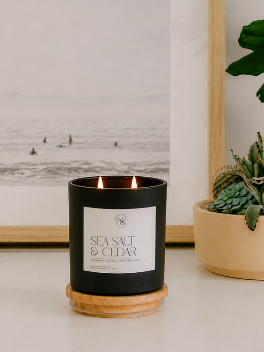 A black candle jar with a white label reading 'SEA SALT & CEDAR' sits on a round wooden coaster on a tabletop. The candle is lit, with two flames visible, and is styled in a home setting next to a potted succulent and a framed black-and-white ocean print.
