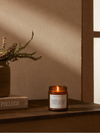 Candle with a label on a wooden surface next to a vase with flowers, against a beige wall.
