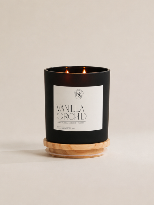 A black double-wick candle in a matte black jar with a white label reading 'VANILLA ORCHID' and a light natural wood base, photographed on a warm off-white background.
