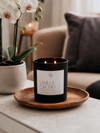 A black double-wick candle labeled 'Vanilla Orchid' sits on a round wooden tray on a light marble table, with a potted orchid and a sofa in the softly blurred background.