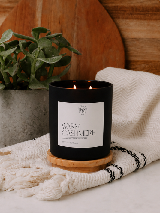 A black candle jar with two lit flames, featuring a white label that reads 'WARM CASHMERE' and resting on a light wood base on top of a folded towel against a warm beige kitchen background.