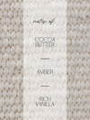 A close-up texture panel with a cream-colored vertical center band reading 'notes of: COCOA BUTTER AMBER RICH VANILLA' in black text, flanked by warm beige and cream marbled knit-like textures.