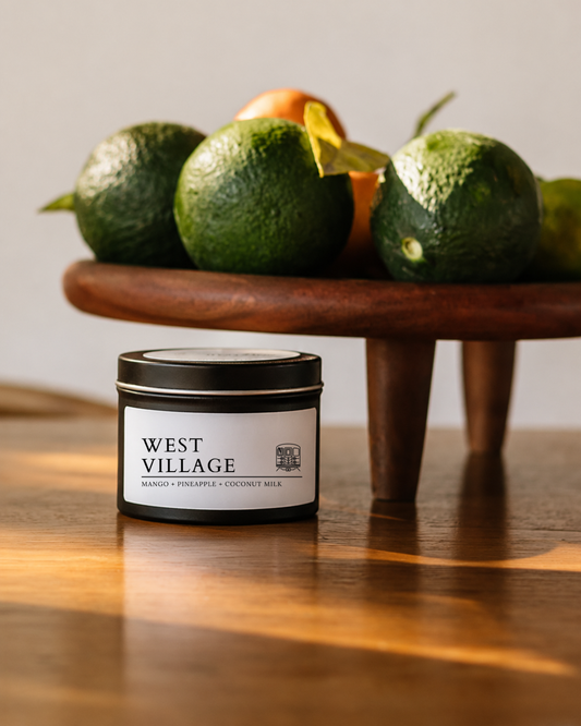 A small black metal candle tin with a white label reading WEST VILLAGE sits on a wooden table in front of a wooden stand holding green avocados and citrus fruits.